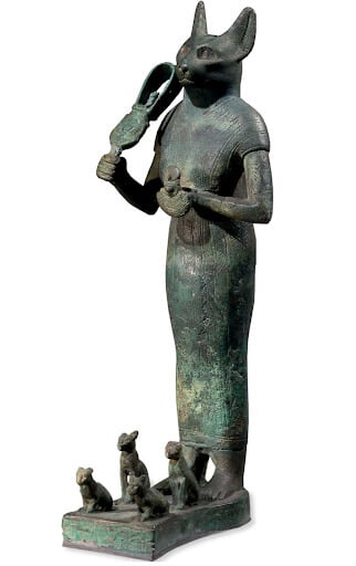 Escultura em bronze da deusa Bastet. Aos pés da deusa estão quatro gatinhos que representam fertilidade. Na mão, ela carrega um instrumento musical que lembra um chocalho. 